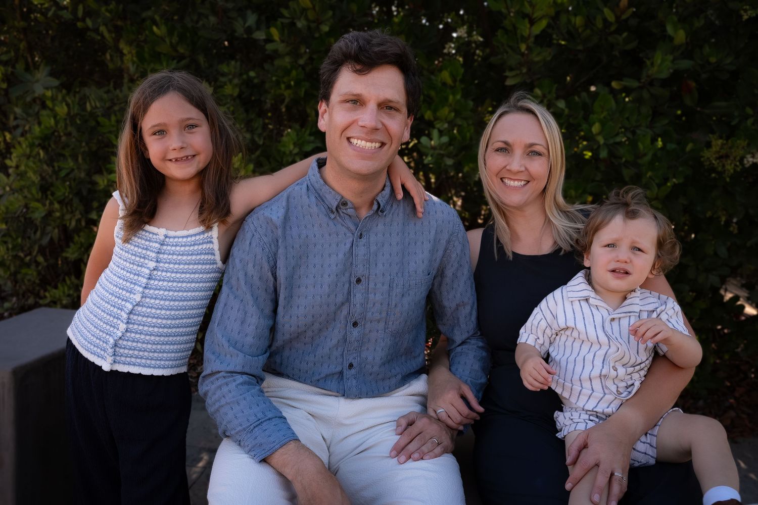 a relaxed family portrait of a father, mother, seven year old daughter and toddler all smiling and looking at the camera