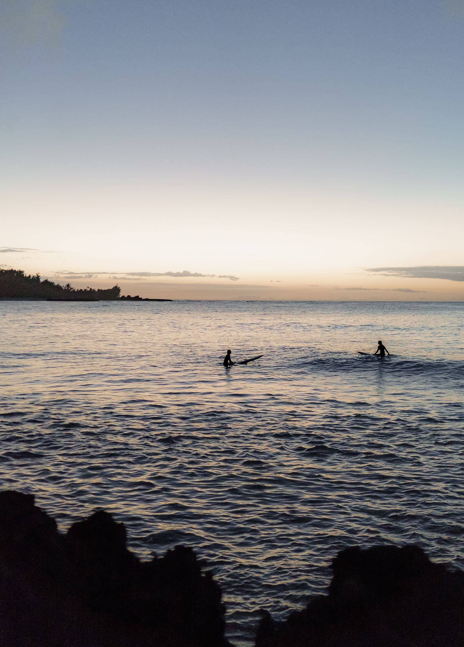 a photo of the ocean surrounded by rocks at sunset with the silhouette of two surfers waiting for a wave