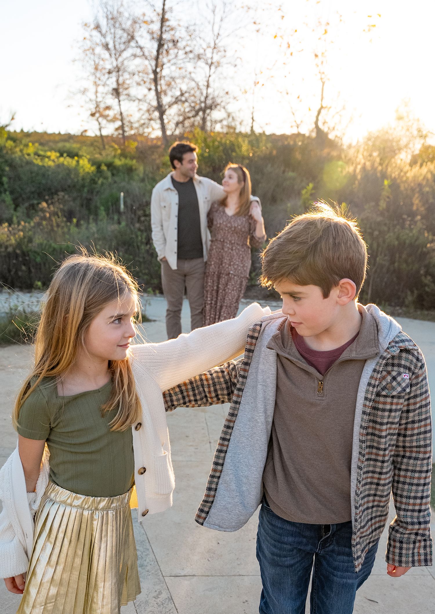 a sister and brother with arms around each other while the parents look on in the background
