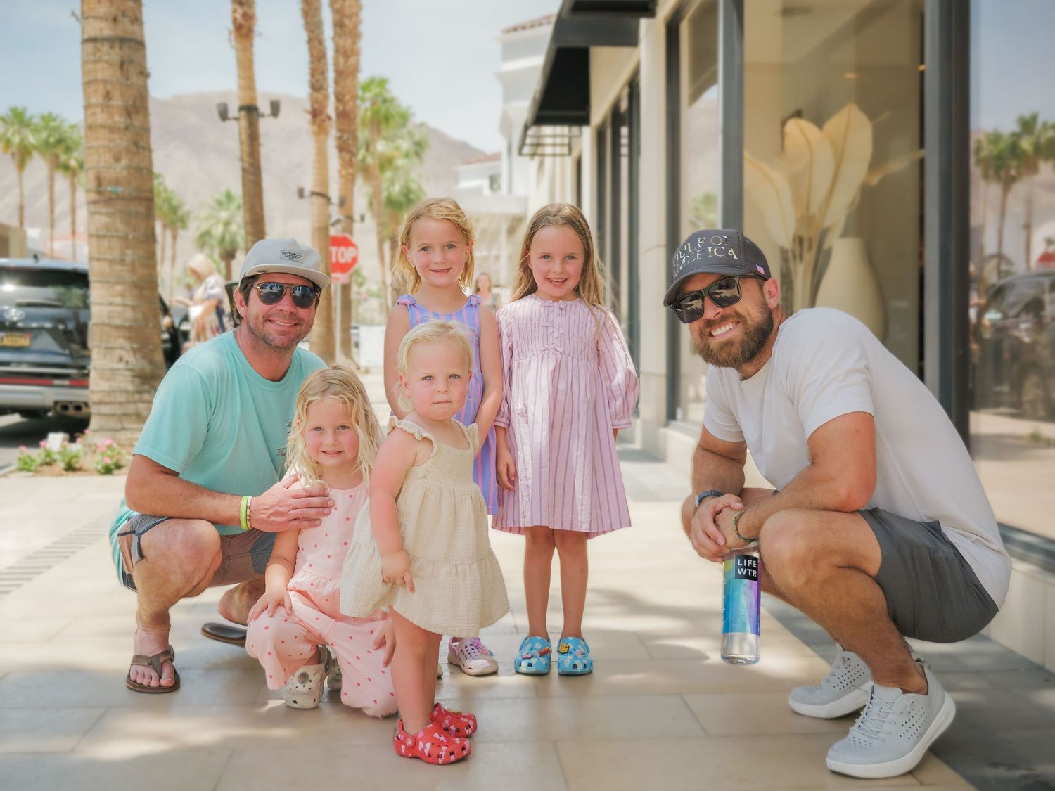 Family members wearing coordinated pink and white outfits pose together on a sunny walkway with palm trees.