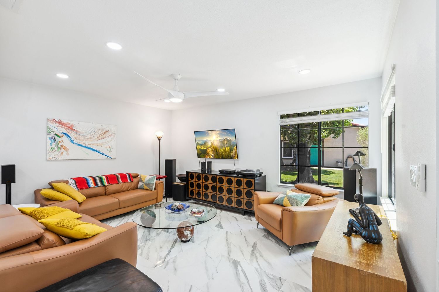 Modern apartment living room with tan leather sofa, wall-mounted TV, and bright natural lighting through large windows.