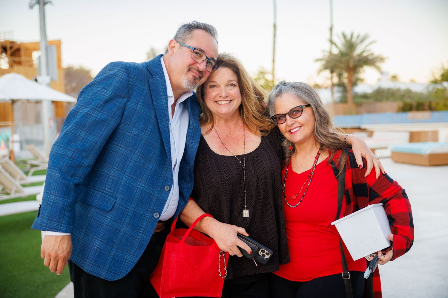 Three people pose together outdoors, with two wearing red and one in a blue blazer at a casual evening event.