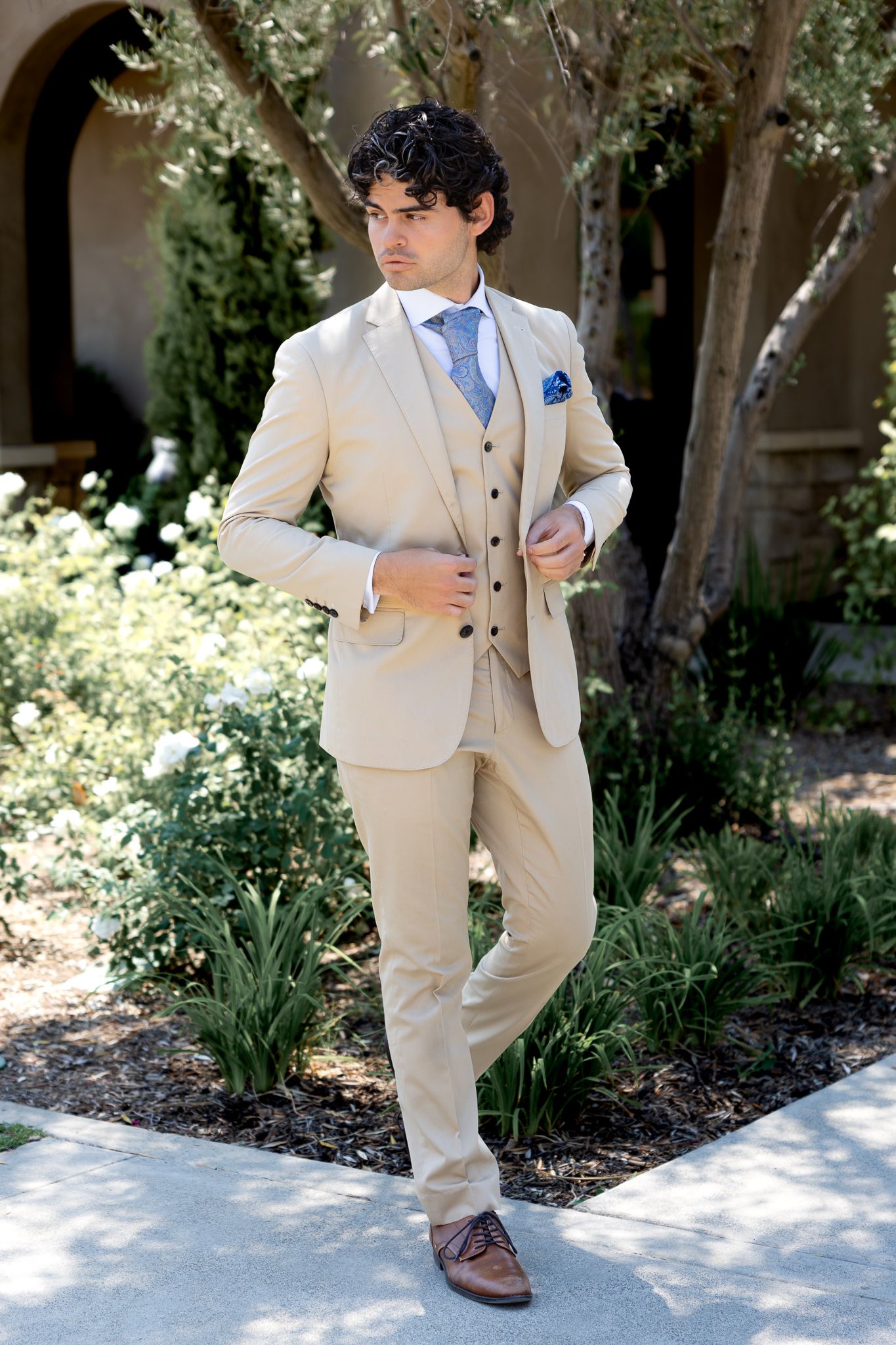 Young Groom in suit strikes playful poses outdoors against a garden backdrop. Editorial posing. Luxury weddings. Cool, edgy groom.