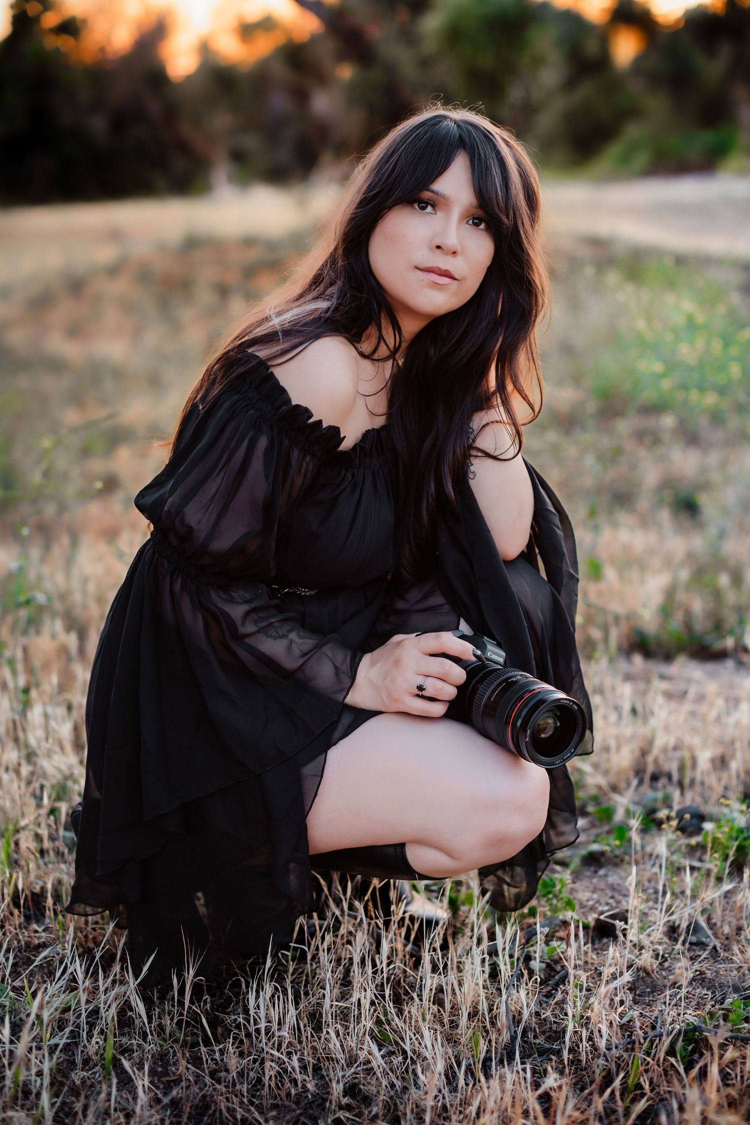 A photographer in a black dress poses with a camera in a golden field during sunset. Moody light Orange County.