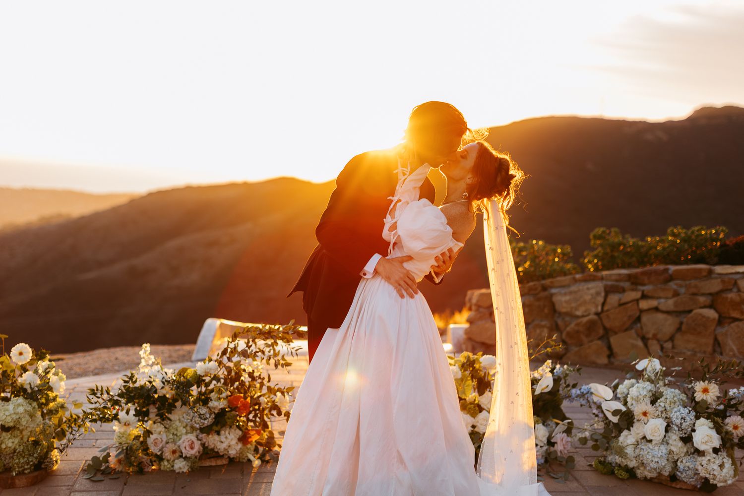 A romantic sunset wedding moment captured at a mountaintop venue with stone walls and white floral decorations.