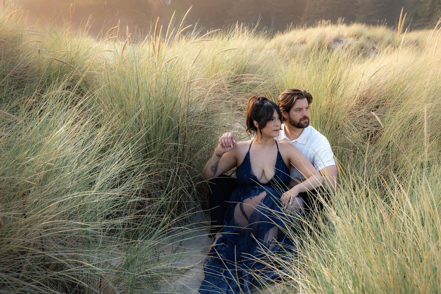 A couple embraces while sitting among tall swaying beach grass during golden hour in Oregon. Canon Beach engagement photos. Engaged couples.