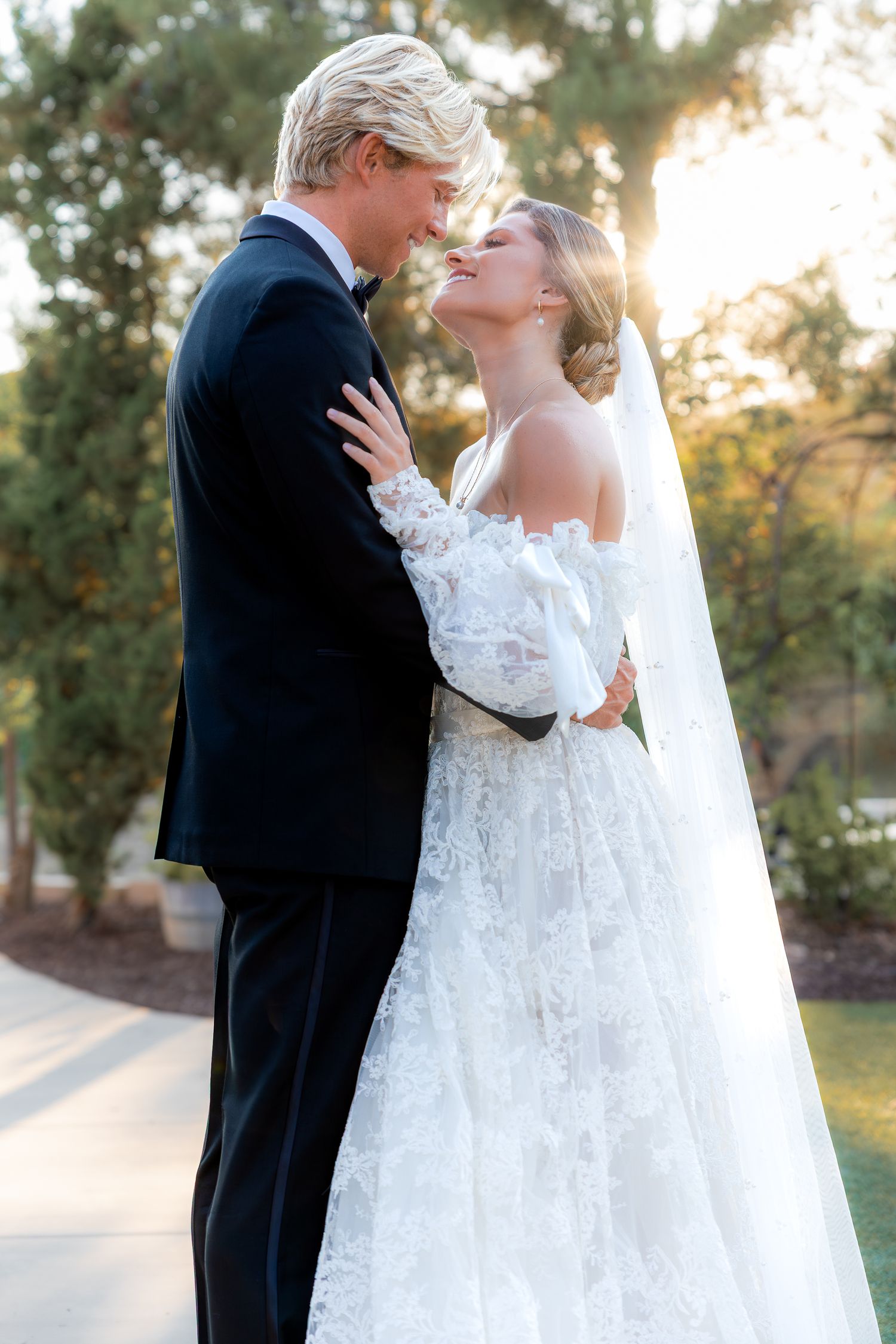 Bride and Groom embrace in a garden during golden hour, with the bride wearing a lace gown and veil. Groom in black tuxedo. Elegant portrait