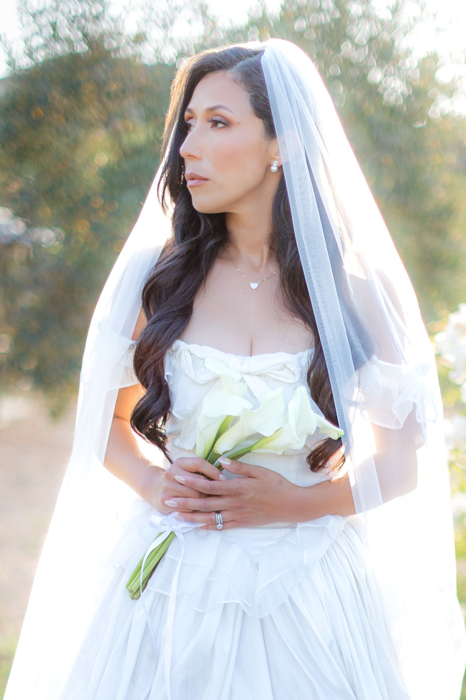 A bride in a flowing white wedding gown and veil poses outdoors during a romantic bridal photoshoot at sunset. Wedding portraits.