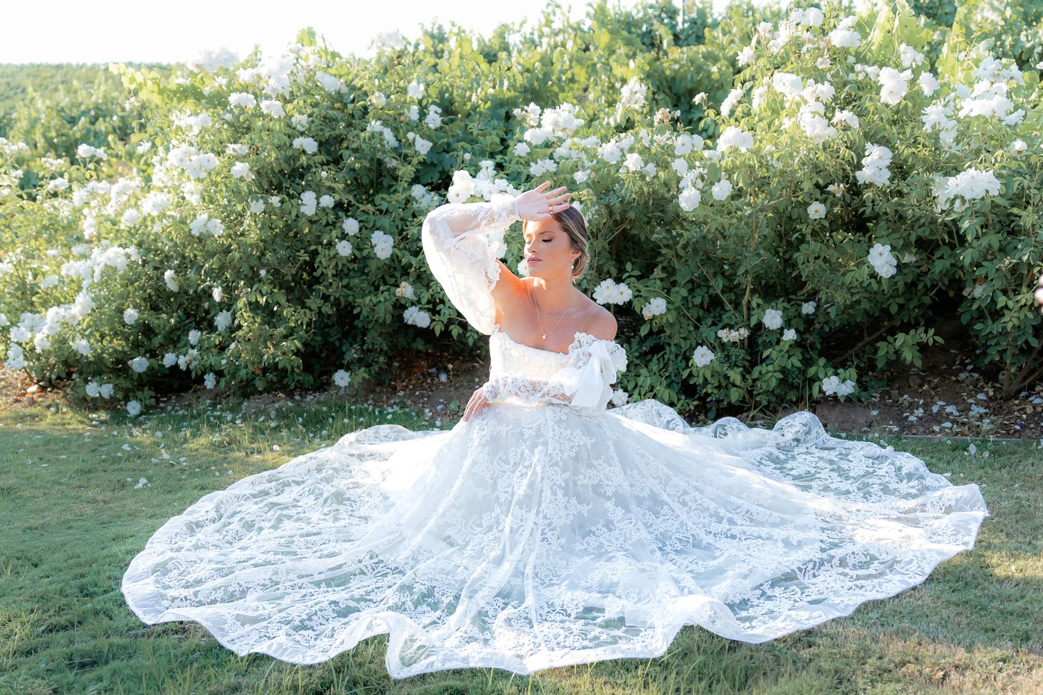 A bride in a flowing white lace wedding dress lies gracefully in a garden during golden hour in Southern California. Luxury photography.