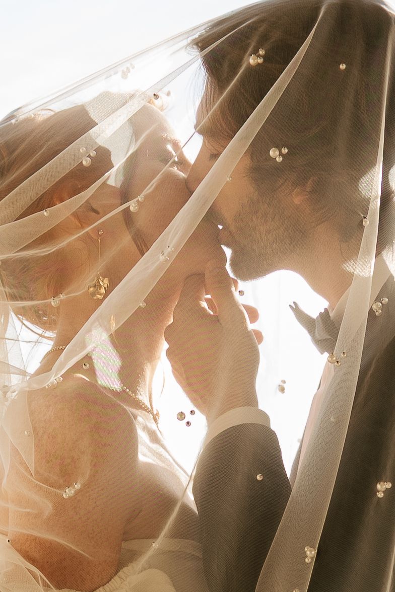 A romantic silhouette of a couple beneath a delicate veil with small floral details, backlit by soft sunlight. Editorial, romantic photo.
