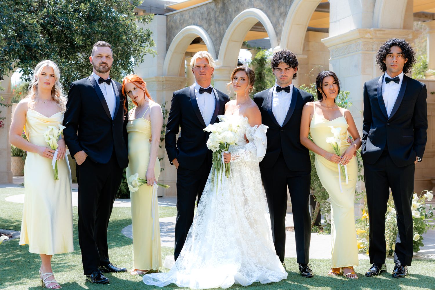 Wedding party in formal attire poses under white stone archways in an elegant outdoor setting. Editorial. Vogue inspired. Elegant photos.