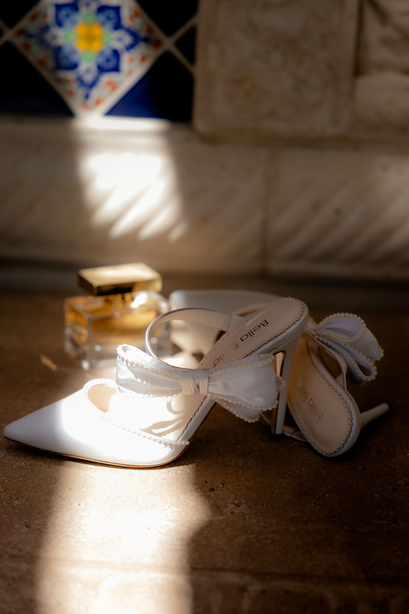 White bridal shoes and perfume bottle illuminated by dramatic sunlight on tiled floor with decorative pattern. Wedding details. High fashion