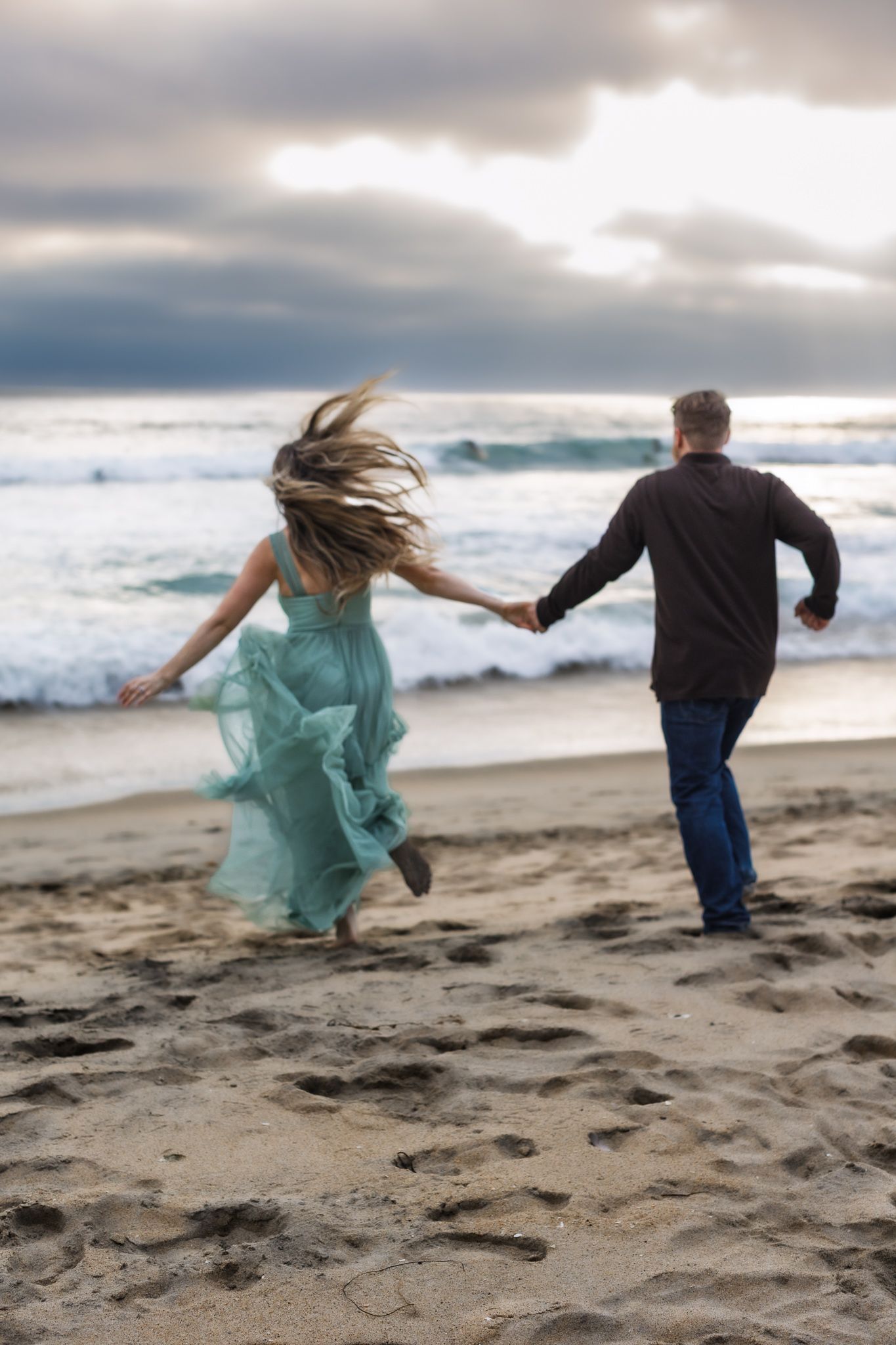 A couple holding hands and running joyfully along a dramatic beach shoreline with crashing waves and cloudy skies. Cinematic. Moody.