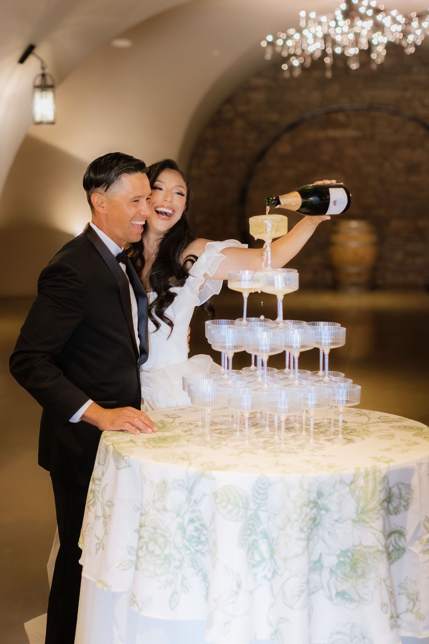 Wedding couple shares a joyful moment while pouring champagne into a tower of glasses at their wedding reception celebration. Luxury venue.