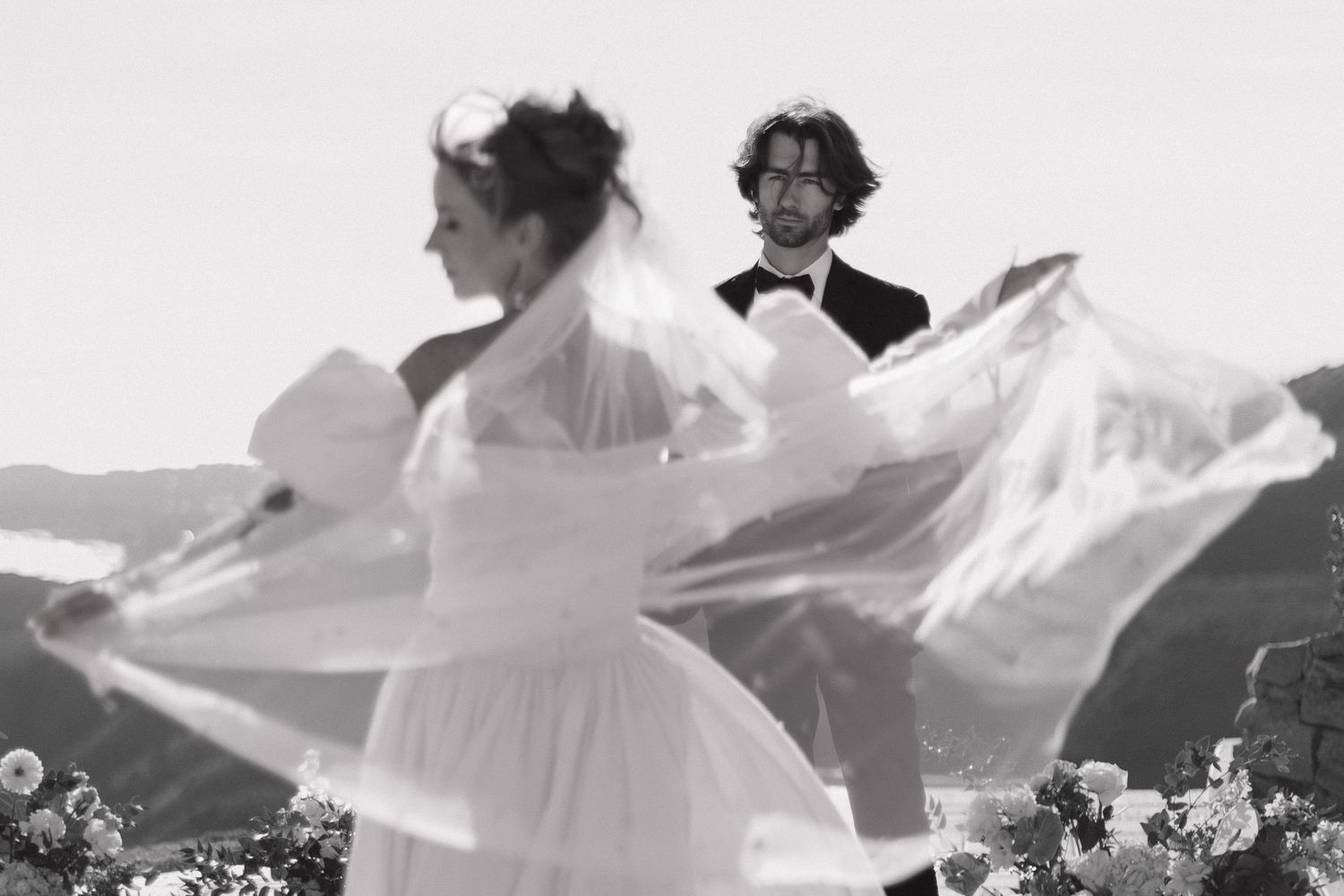 A flowing white wedding dress billows in the wind against a scenic mountain landscape backdrop during wedding portraits of bride and groom.