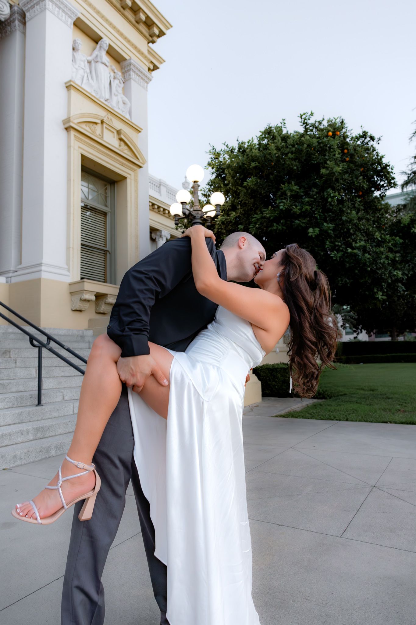 Engaged couple in formal attire share a passionate dip kiss on stone steps outside beautiful architecture. Fun and flirty engagement photos.