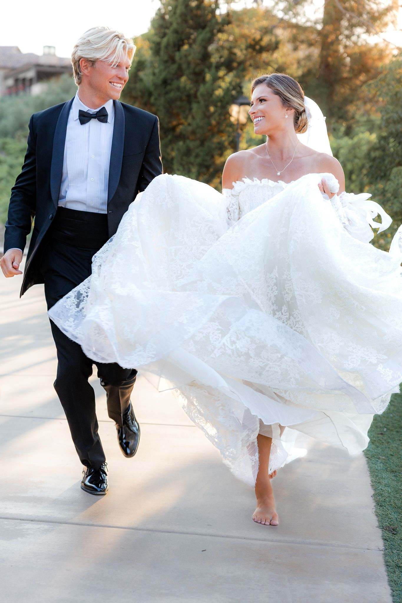A couple walks together outdoors on their wedding day, with the bride's white lace gown flowing in the breeze. Happy bride and groom photos.