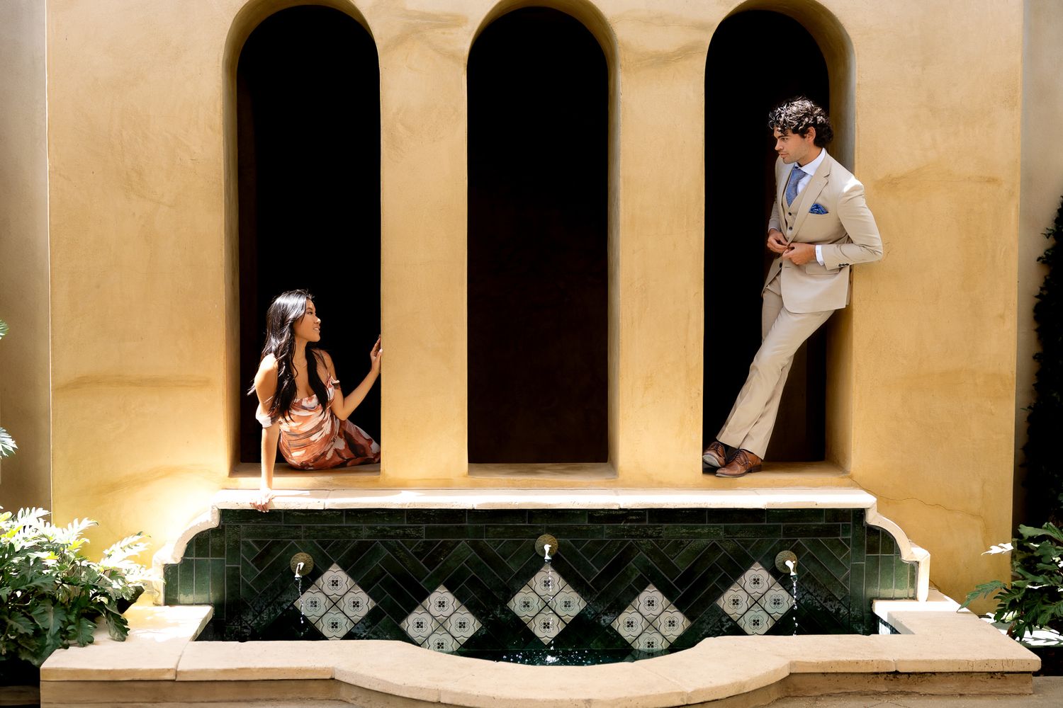 An engaged couple pose in archways of a Mediterranean-style building with decorative tile work and beige walls. Luxury engagement photos.