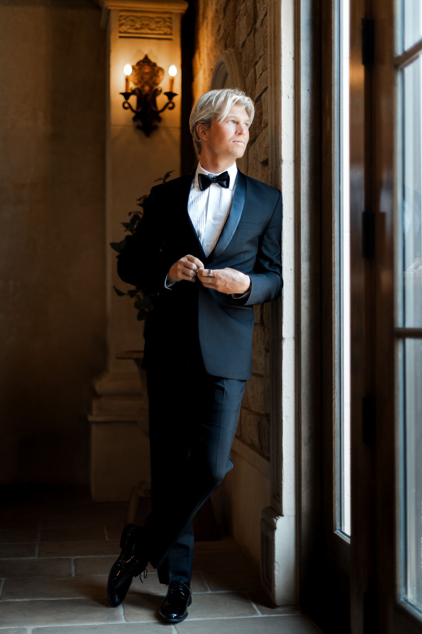 A man in a tuxedo leans against a window in an elegant hallway with ornate architectural details.