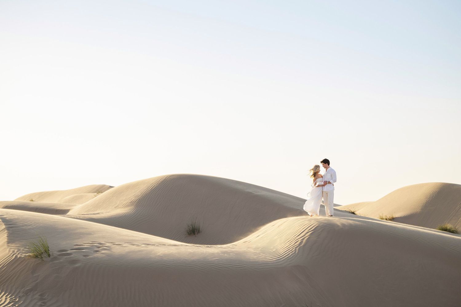 Two people stand embracing on rolling sand dunes in soft desert light during a romantic photo session.
