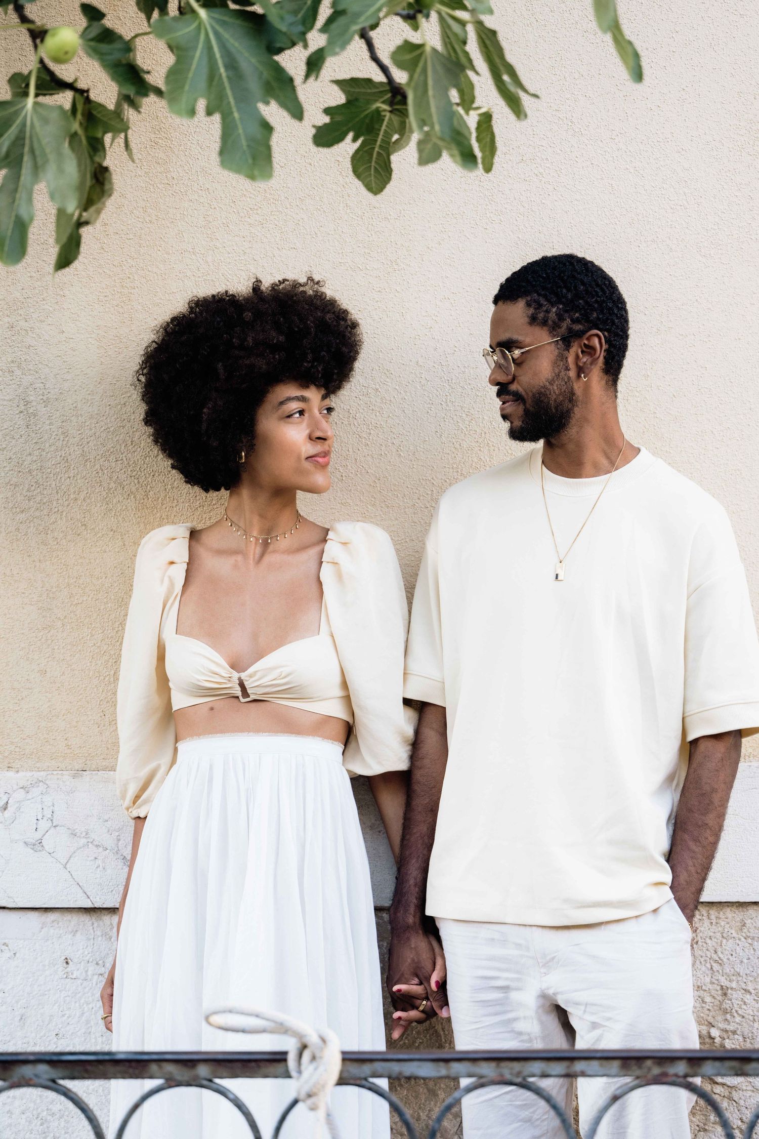 Couple dressed in white shares candid moments against neutral wall with greenery.