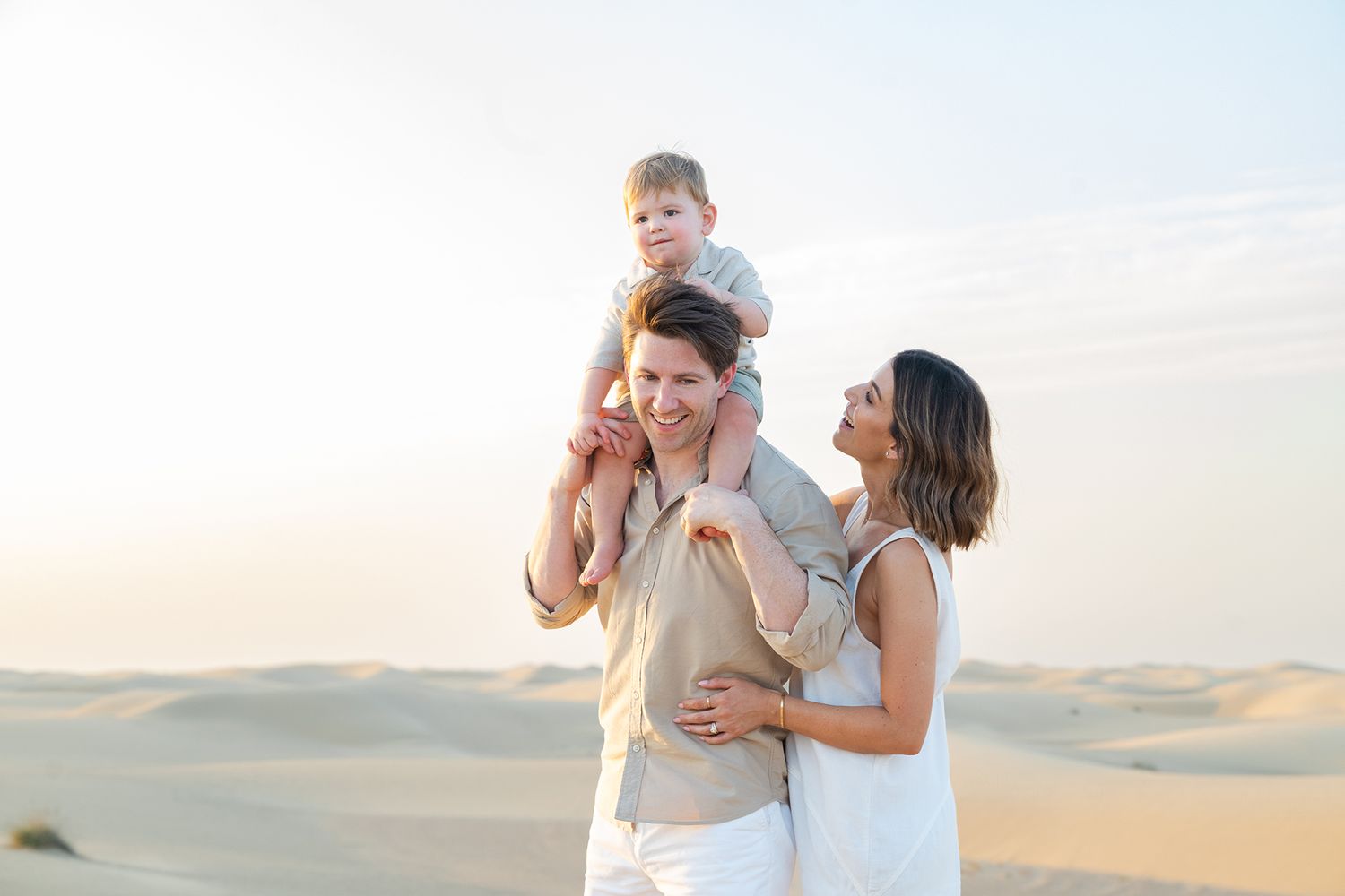 A happy family of three poses on sandy desert dunes, father carrying toddler on shoulders with mother beside them.