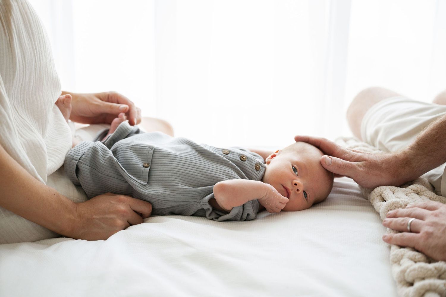 Baby in gray onesie lies on white bedding while parent's hands gently touch their feet and head.