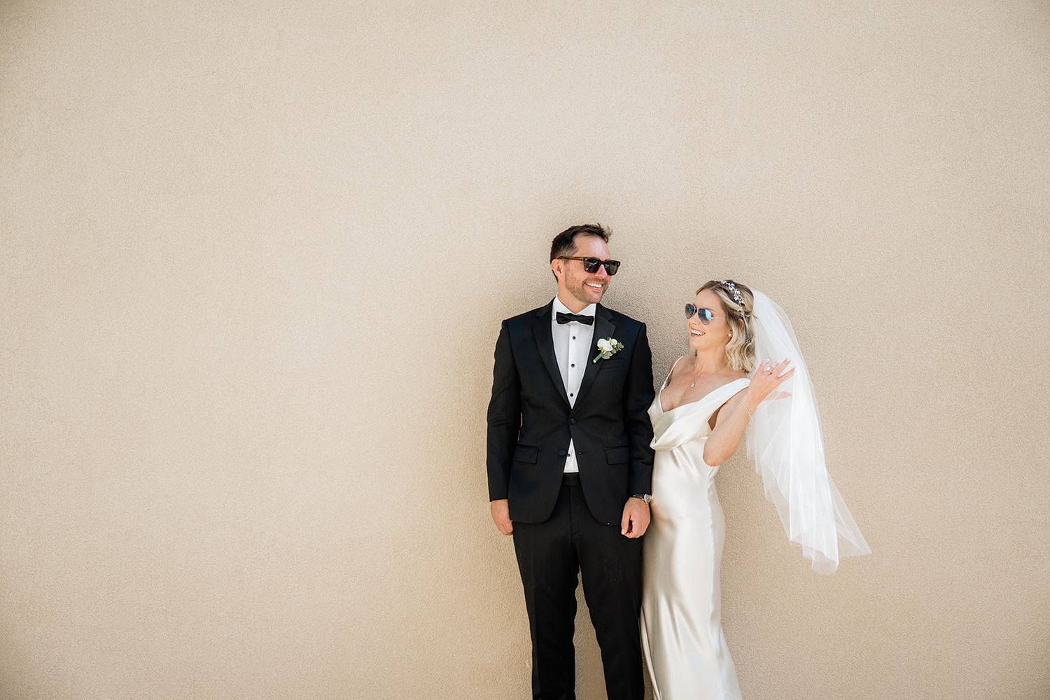 Wedding couple posing playfully against neutral wall in formal attire.
