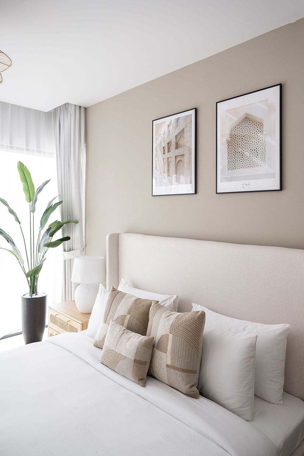 Modern bedroom interior with white bedding, beige walls, framed artwork and potted plants creating a serene atmosphere.