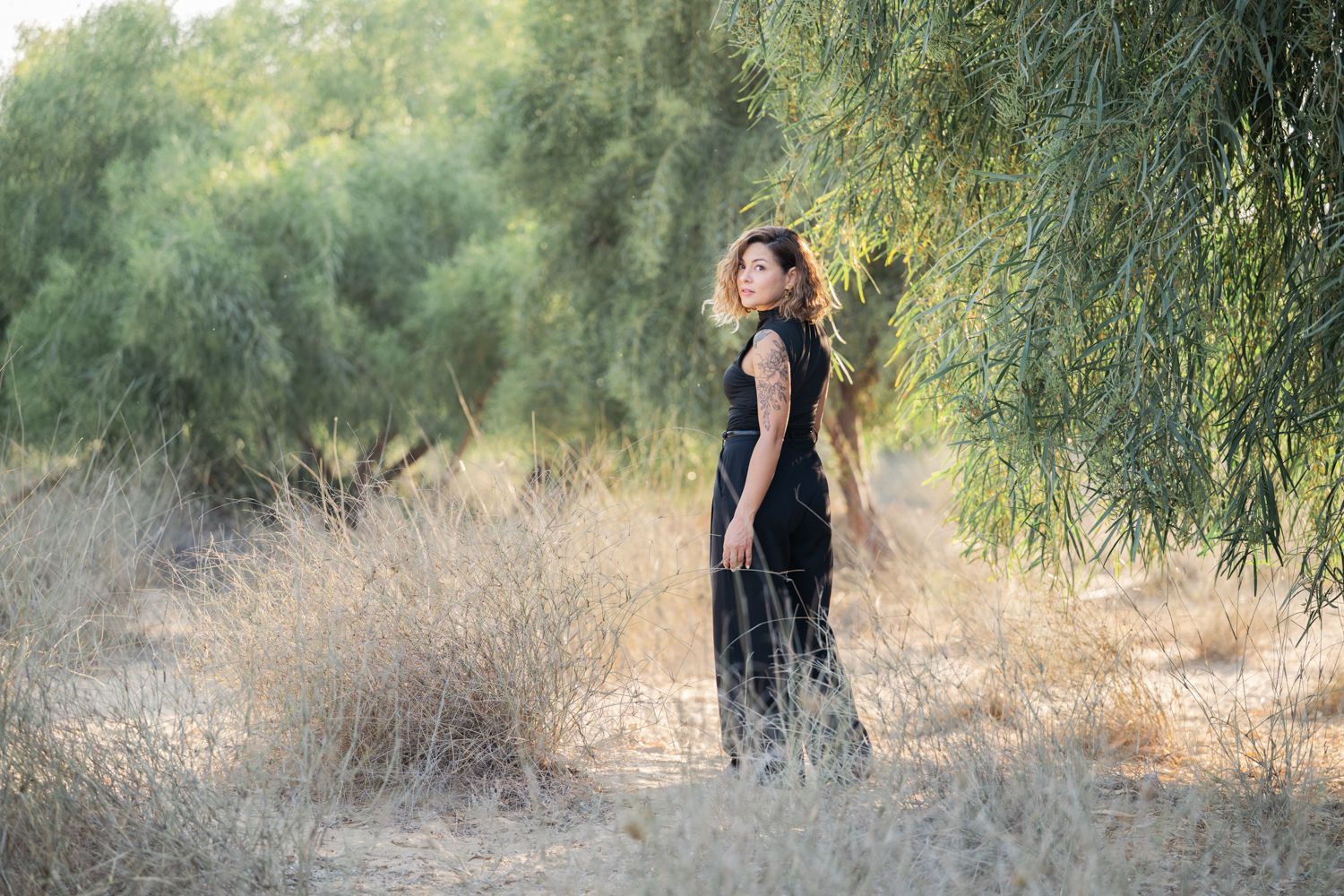 A lady stands in tall grass beneath a tall green tree in soft golden sunlight.