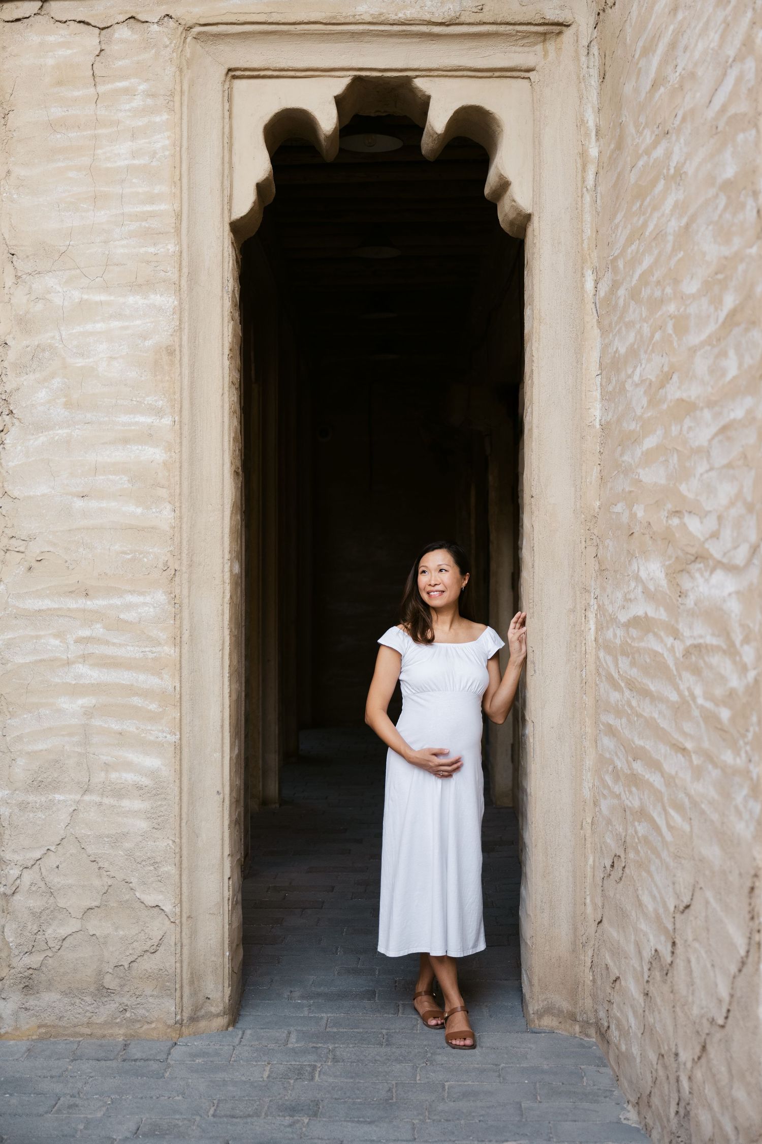 Expectant mother in white dress poses in archways of traditional Middle Eastern architecture.