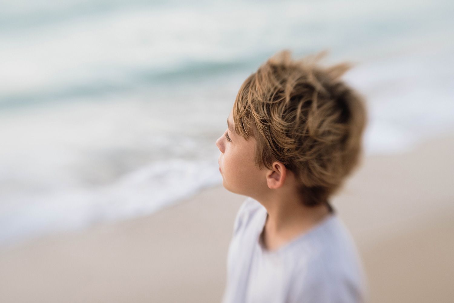 Someone with windswept hair gazes at the ocean from a sandy beach in soft dreamy lighting.