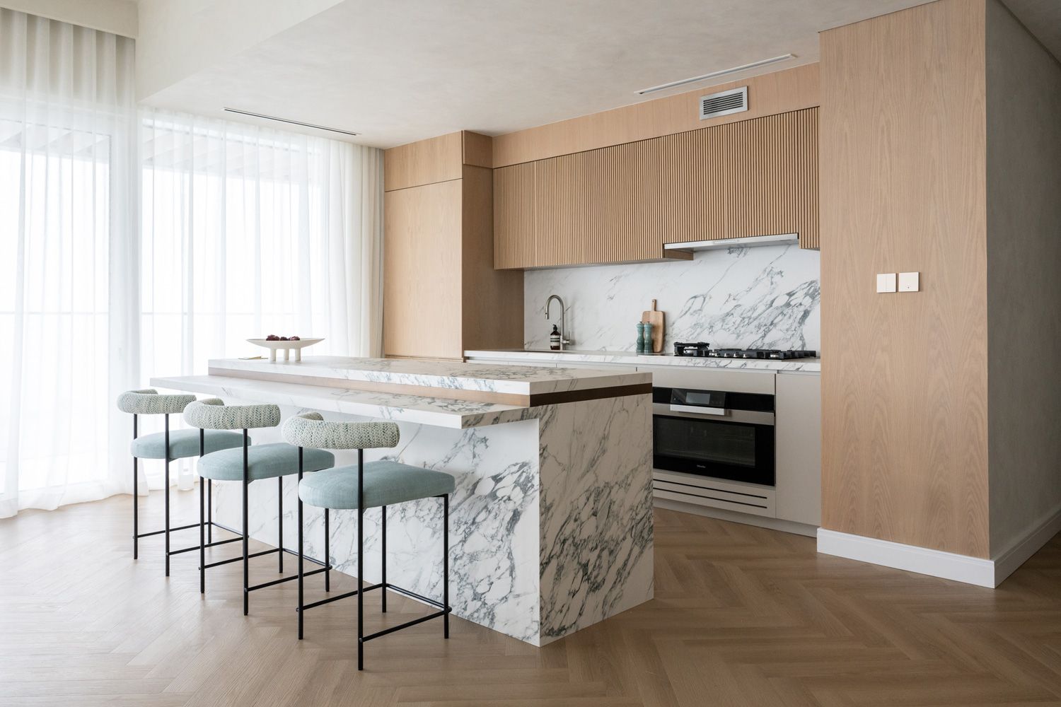 Modern kitchen interior with marble waterfall island, mint green barstools, and light wood cabinetry on herringbone floors.