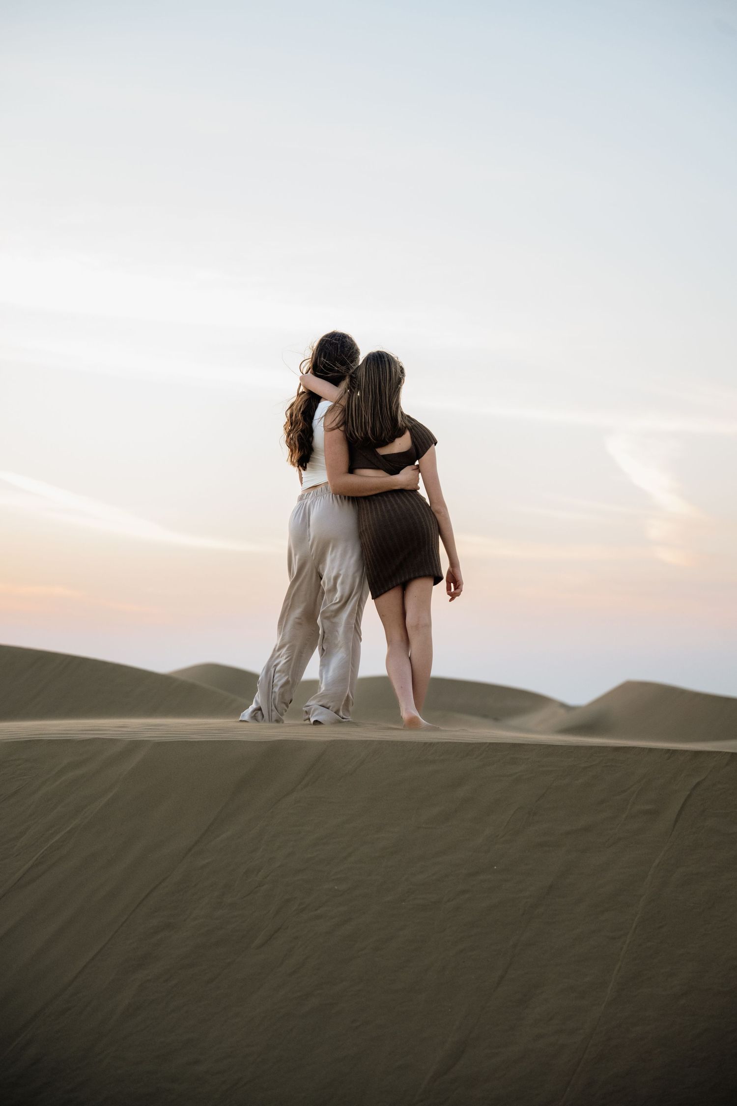 Couple embracing while sitting atop desert sand dunes during sunset.