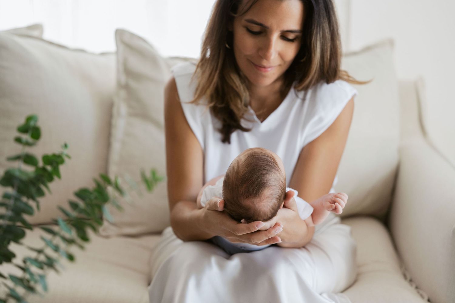 Mother and newborn baby share tender moments together while sitting on a white couch at home.