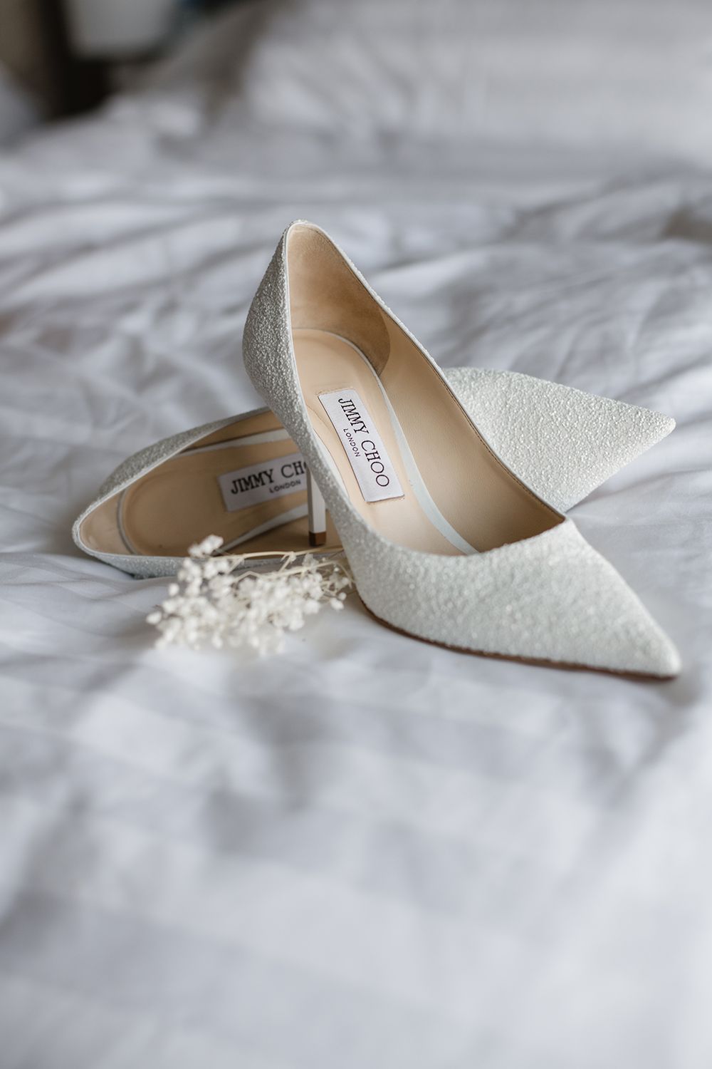 Elegant white lace Jimmy Choo bridal shoes on light bedding.