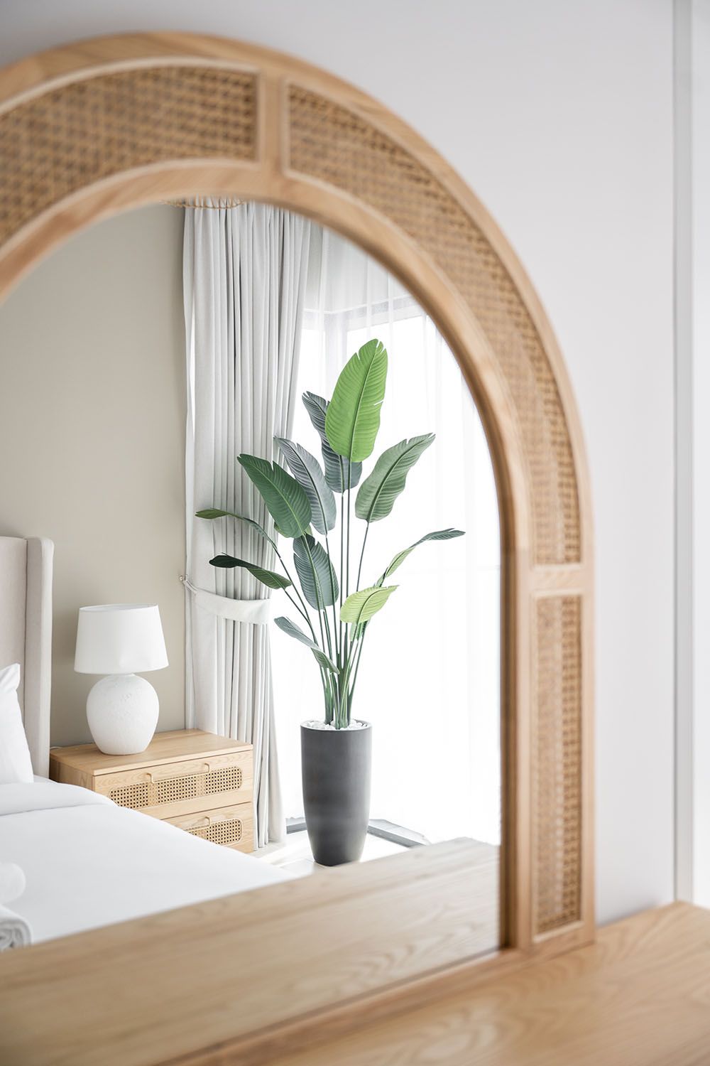 Large arched rattan mirror reflecting a bright interior space with tropical plants and white curtains.