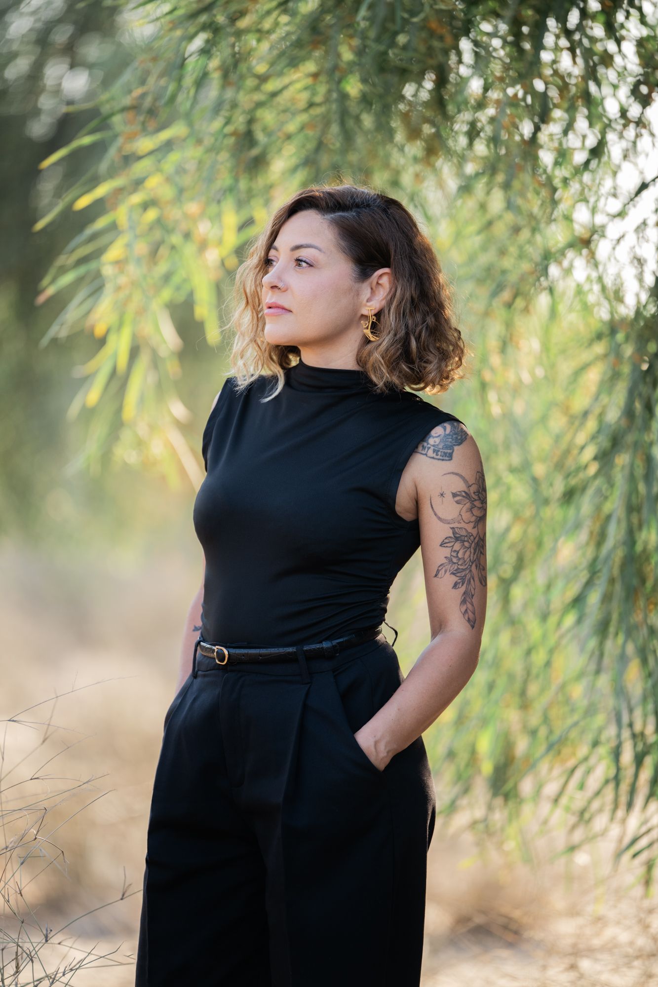 A lady in a black dress poses gracefully under drooping tree branches in a natural outdoor setting.