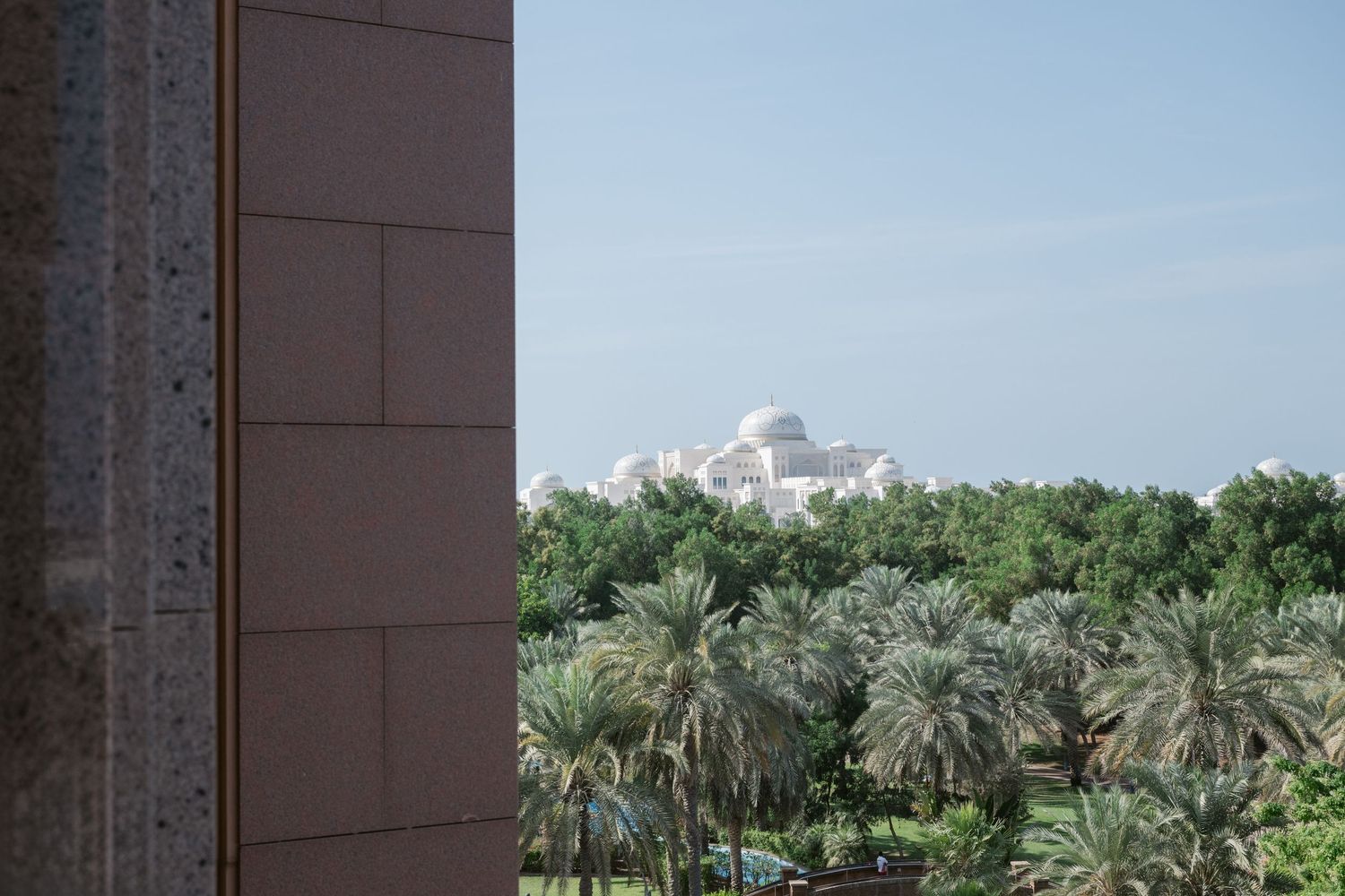 Palm trees line a path leading to a grand building with domed architecture in a sunny desert landscape.