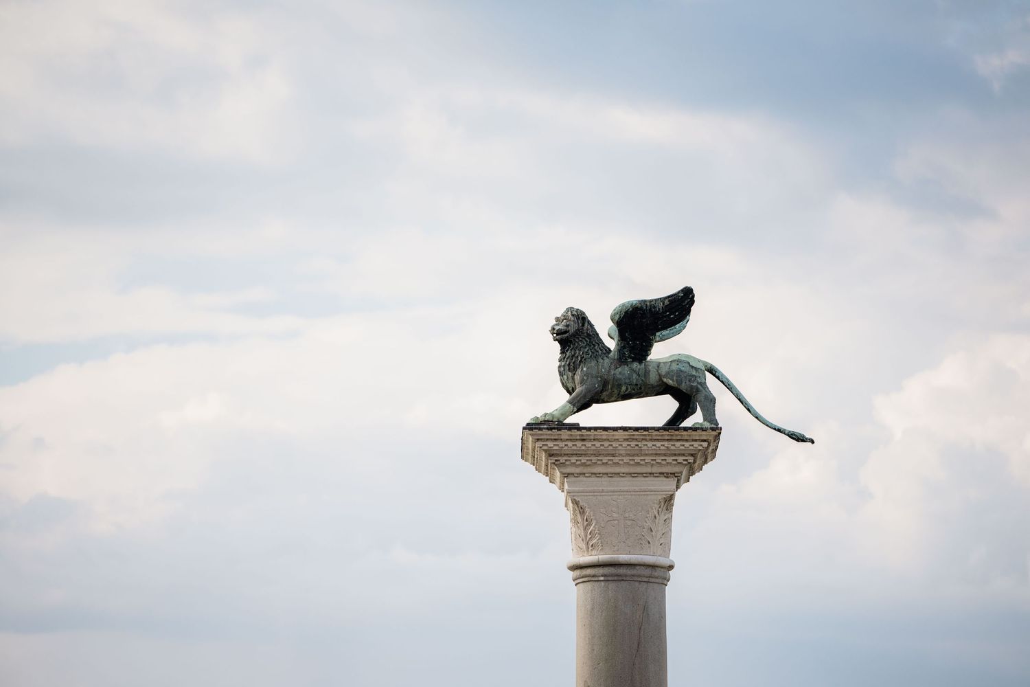 Winged lion statue atop an ornate stone column against a cloudy sky in Venice.