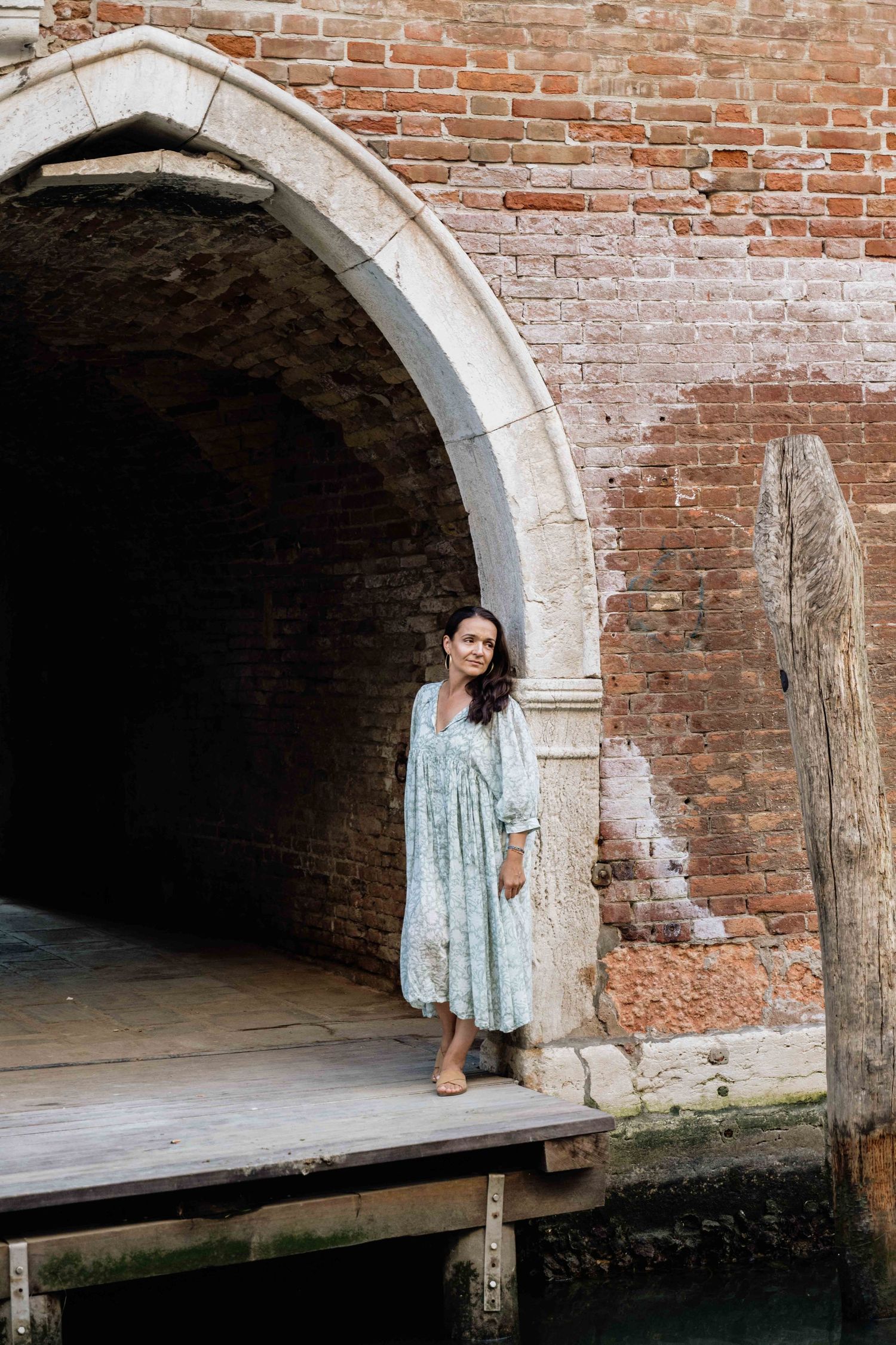 Model in light dress poses on wooden platform by Venetian canal and brick archway.