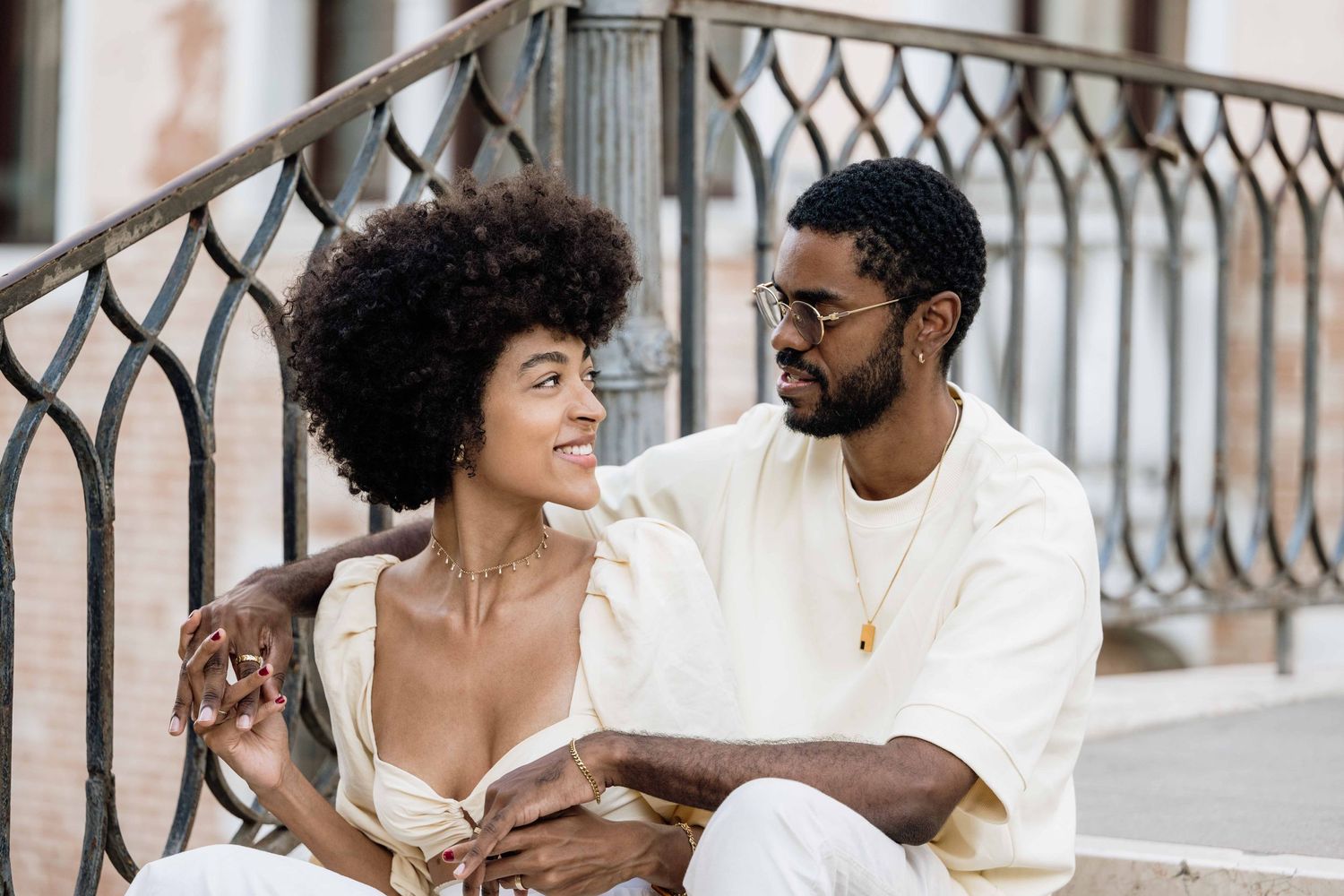 Couple in white clothing poses affectionately on vintage balcony railing.