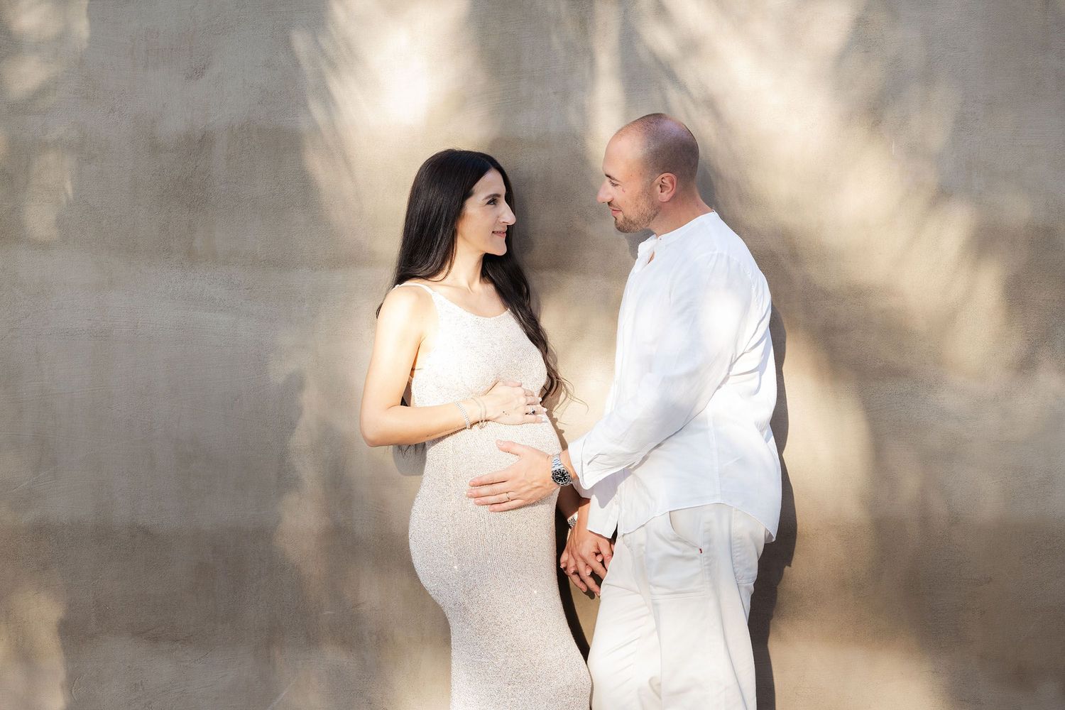 A couple in white outfits shares an intimate moment against a textured wall with dramatic lighting.