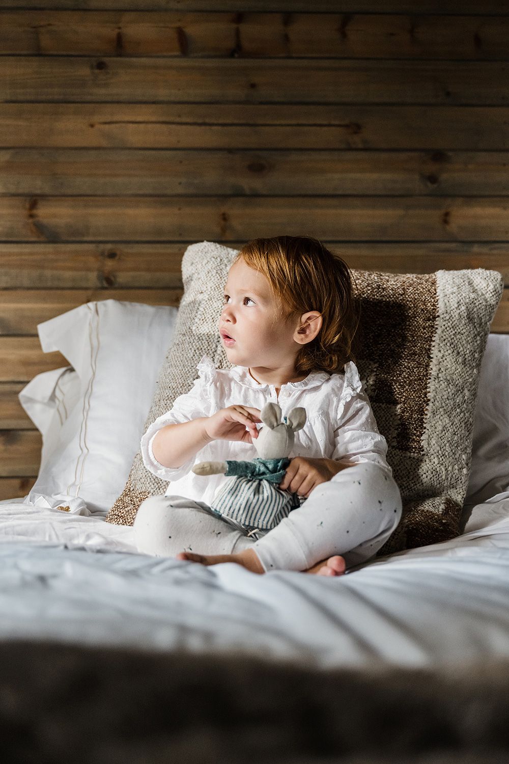 A small figure rests on white bedding against a rustic wooden wall backdrop in a cozy bedroom setting.