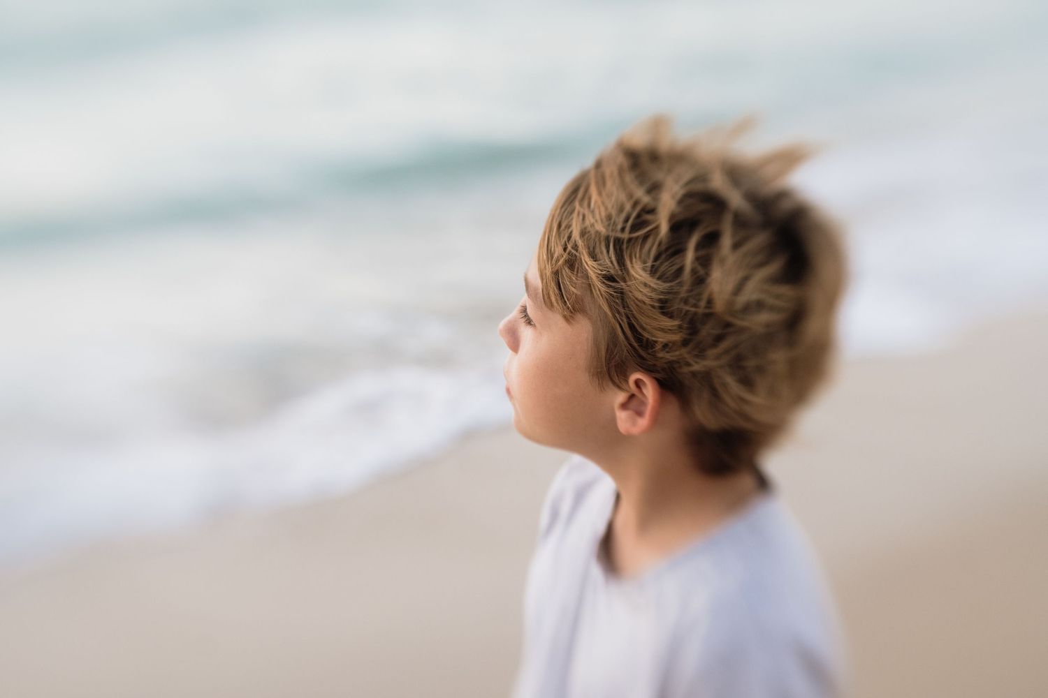 Boy with windswept hair enjoying a peaceful moment on a sunny beach with ocean waves in the background.