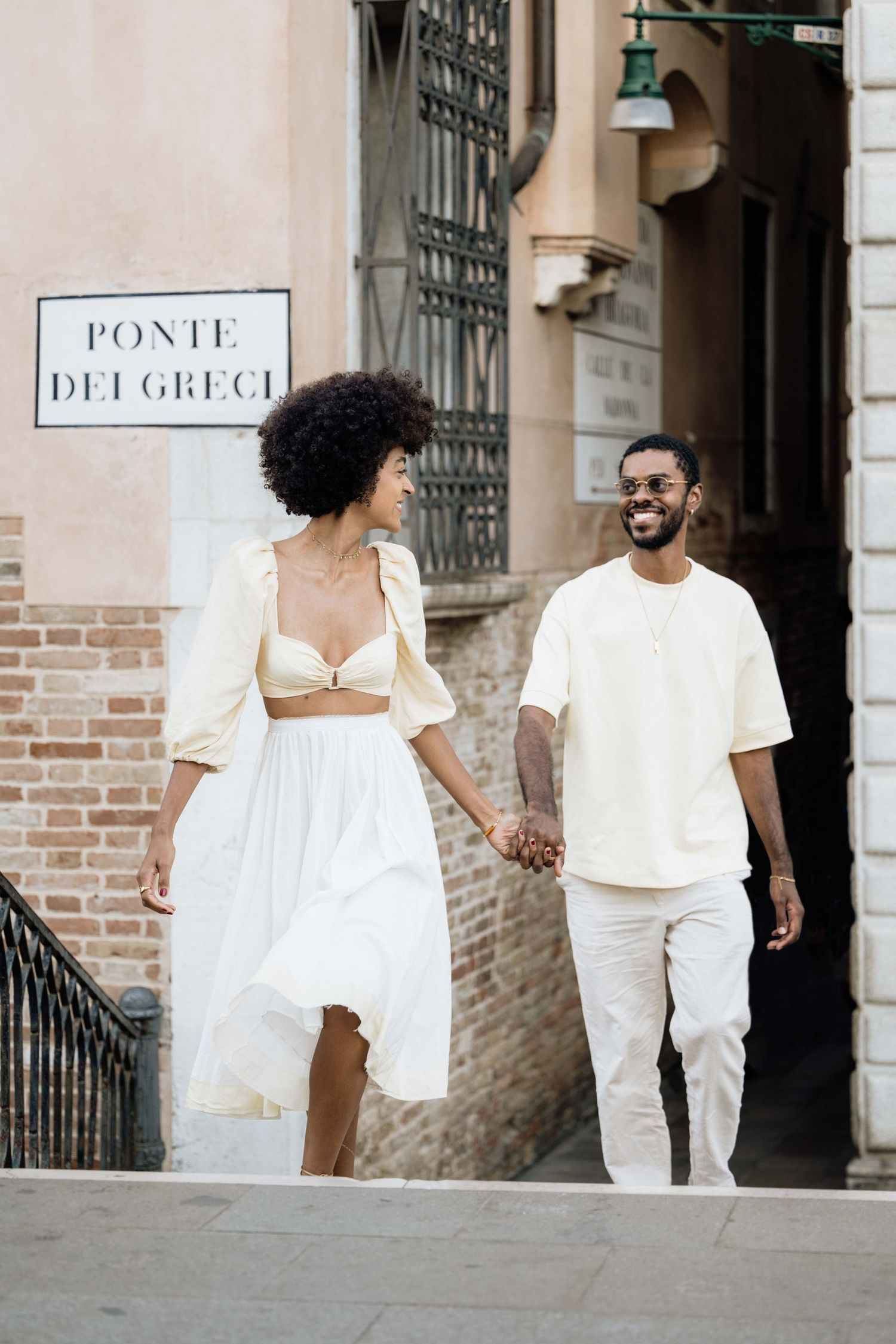 Romantic couple dressed in white walking together along a historic building facade.