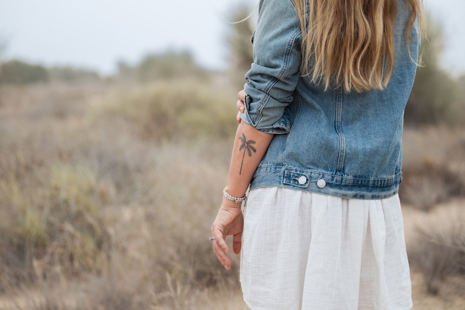 Person in denim shirt and white skirt stands in desert landscape with muted earth tones and dry brush.