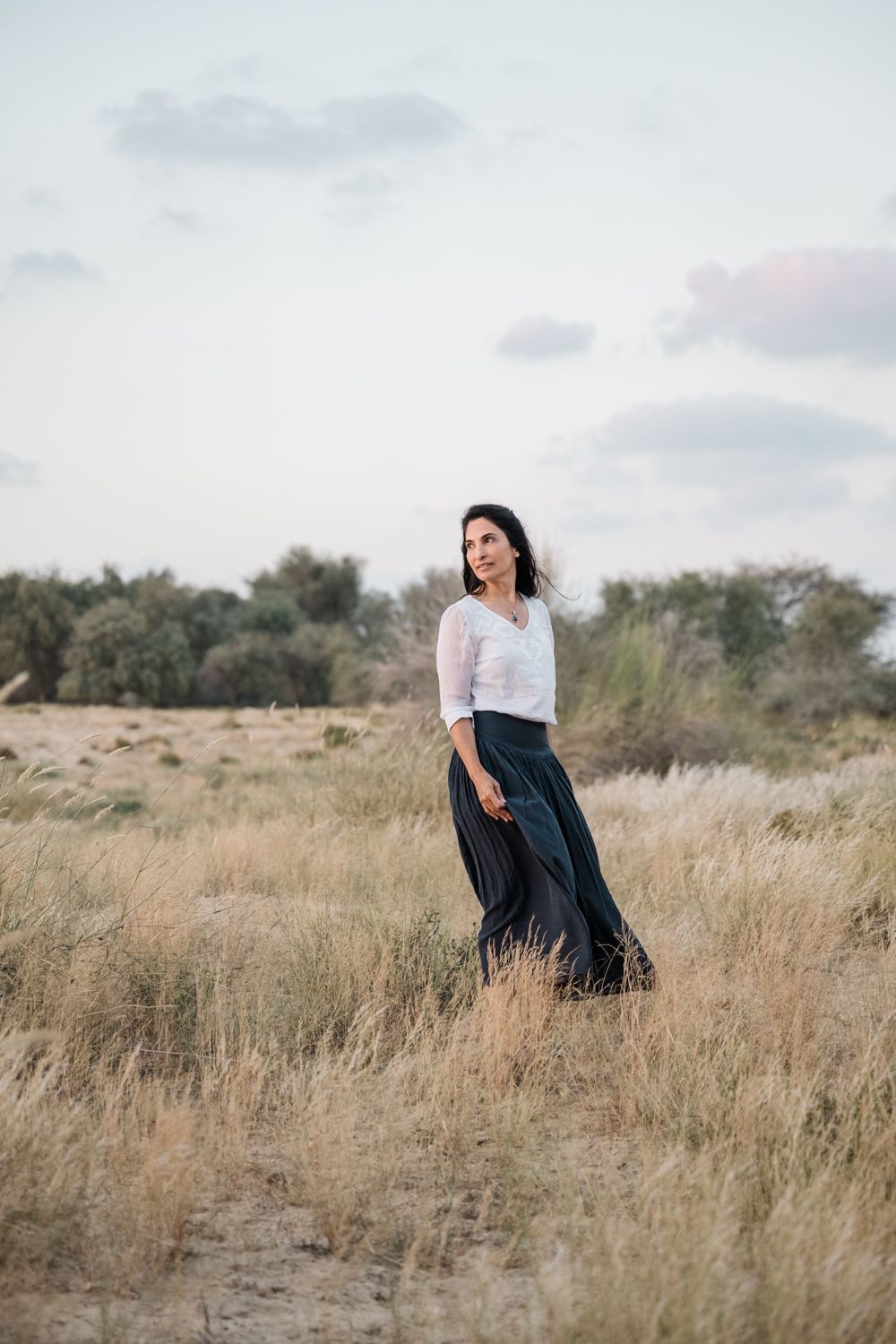 Person in white top and flowing dark skirt poses in golden field at sunset.