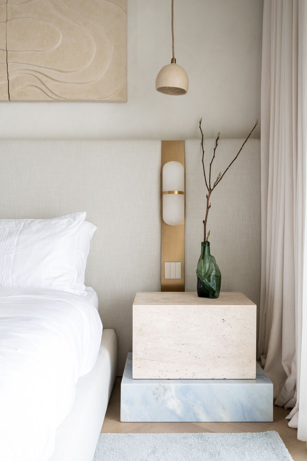 Minimalist bedroom interior with white bedding and green glass vase on light wood nightstand against neutral wall.