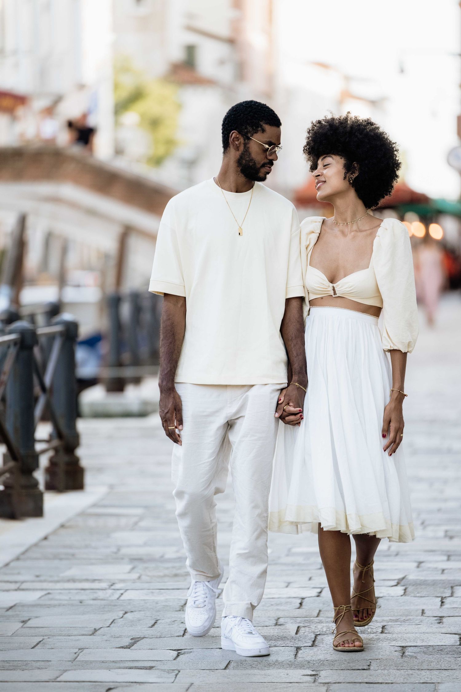 A couple takes a romantic stroll together on cobblestone streets wearing all-white outfits.